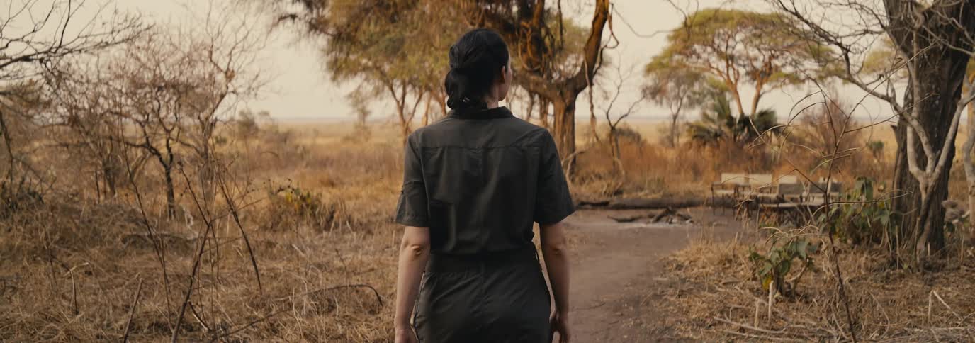 A woman walks alone through dry sunlit grassland toward a rustic tree covered clearing
