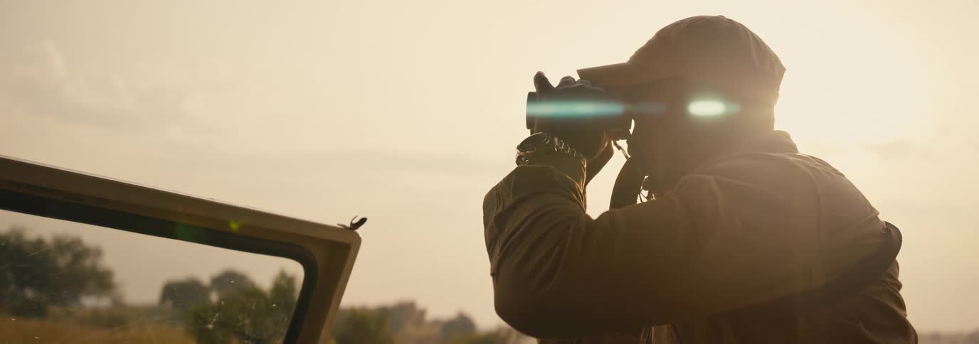 A person is looking through binoculars while standing on an open car door during sunset