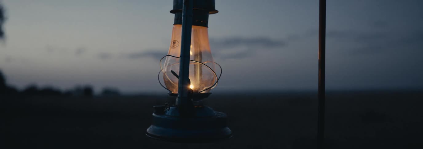 A glowing lantern hangs in the twilight over a dark field with faint tree silhouettes lining the horizon
