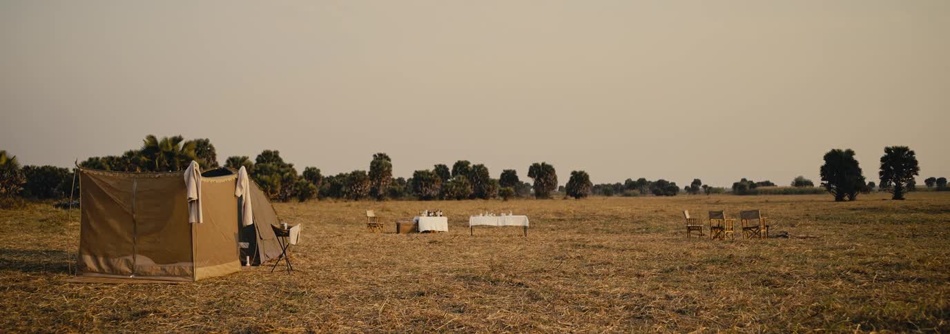 A campsite in a dry field features a tent a long dining table covered with white cloths and several chairs bordered by sparse trees