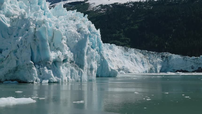 A massive glacier meets calm water with forested mountains behind