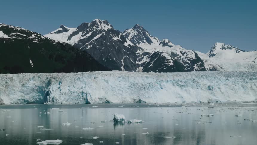 Aerial of Alaska with massive glacier stretches across a calm bay with snow capped mountains in the background