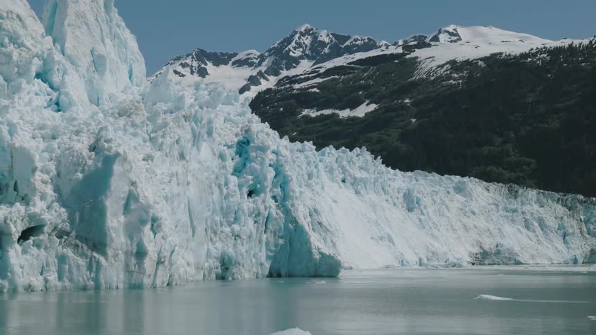 A glacier meets still water beneath snowy mountains and clear skies