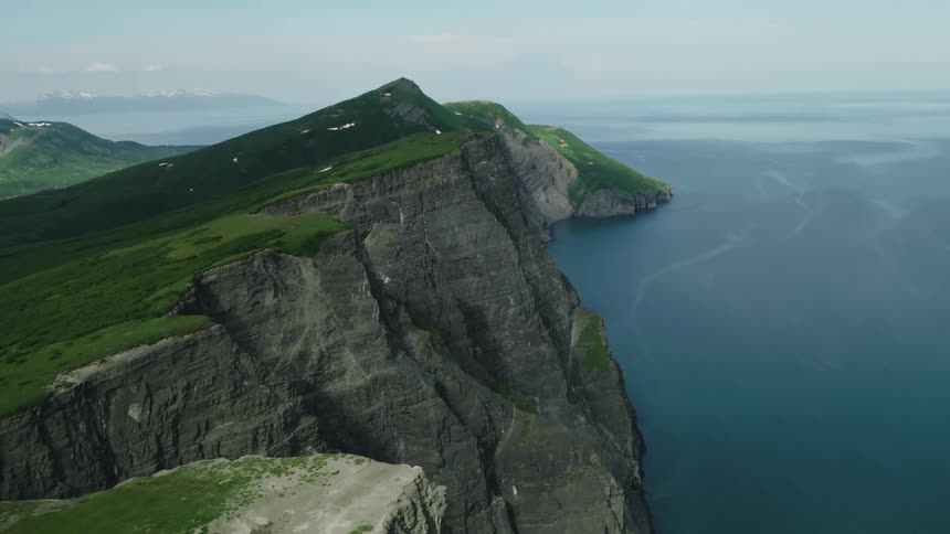 A rugged cliffside covered in lush greenery overlooks a calm blue ocean under a clear sky