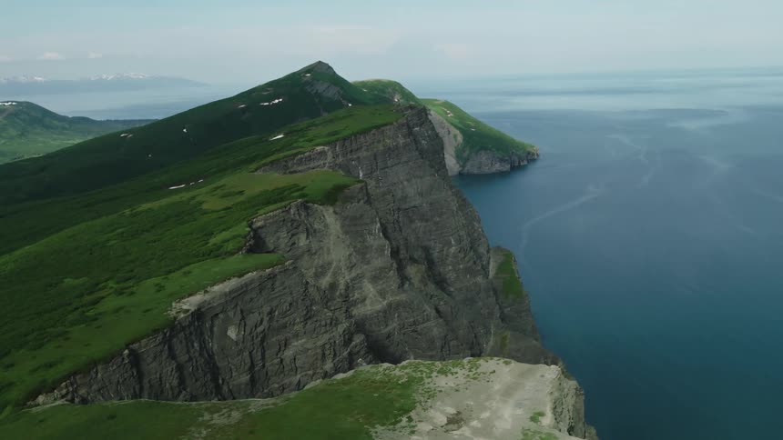 An aerial view of Alaska with huge cliffs covered in green grass facing a blue ocean
