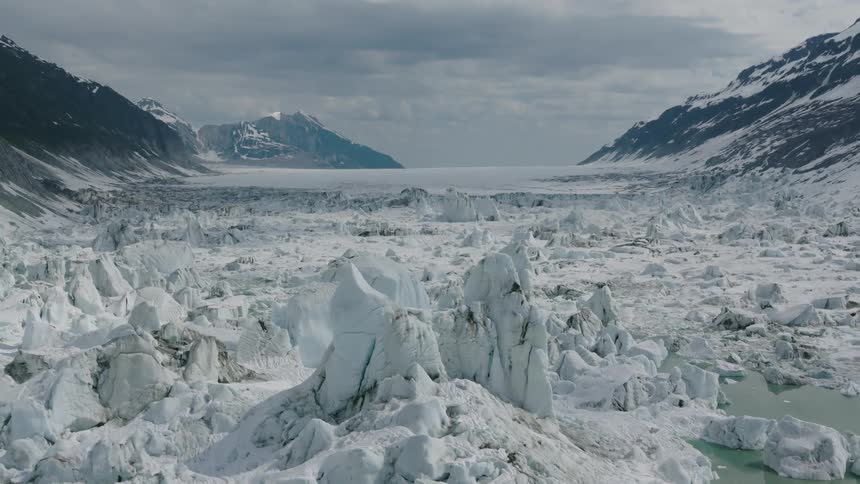 A vast glacier field stretches between snow covered mountains under a cloudy sky