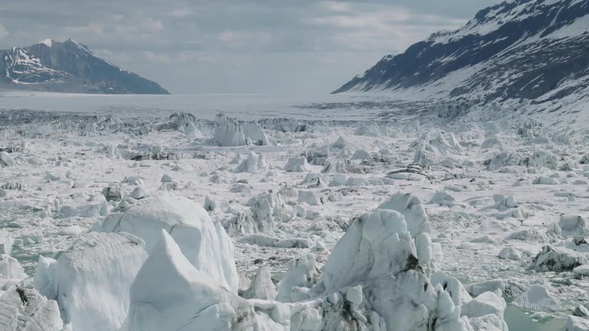 Jagged ice stretches between snowy mountains under a cloudy sky