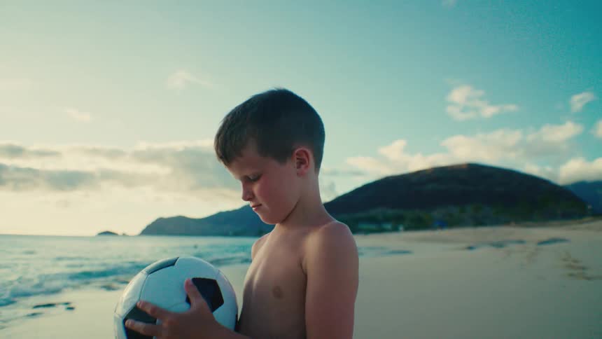 Boy stands on the beach thinking as he holds a soccer ball