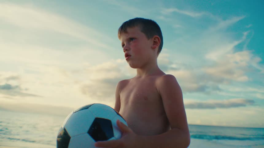 Boy stands on the beach thinking as he holds a soccer ball