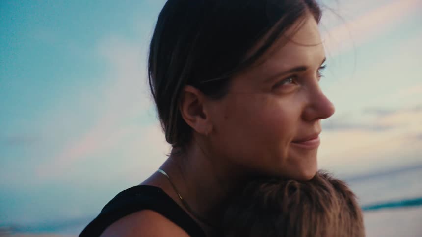 Close up of a mother hugging and kissing her son as they sit outdoors at the beach during daytime