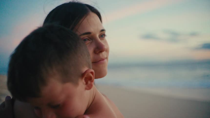 An adult carries a child along a sandy beach at sunset with waves rolling in beneath a vivid multicolored sky