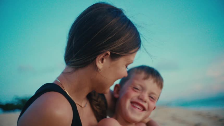 A smiling mother hugs her young son on a sunny beach