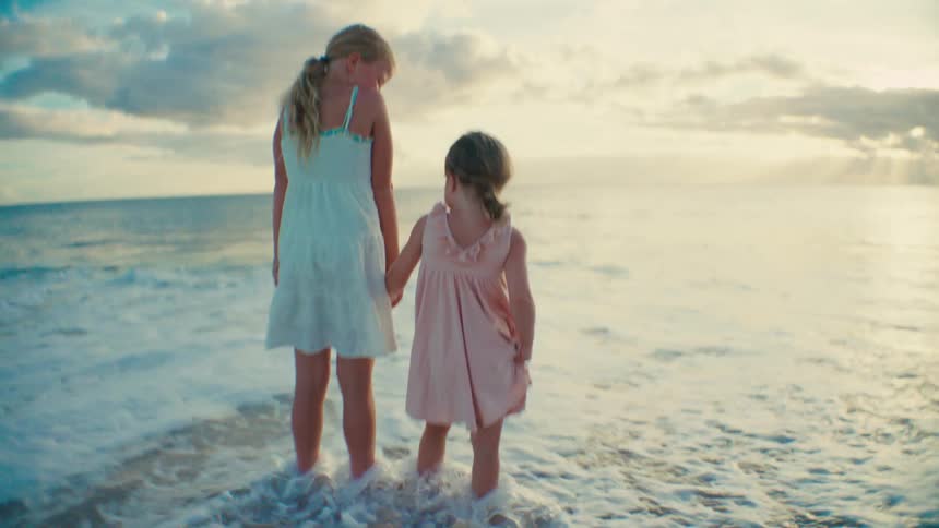 Sisters stand holding hands at the beach enjoying waves at their feet