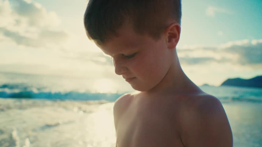 The face of a little boy who is on a beach and he is looking at something that is below him