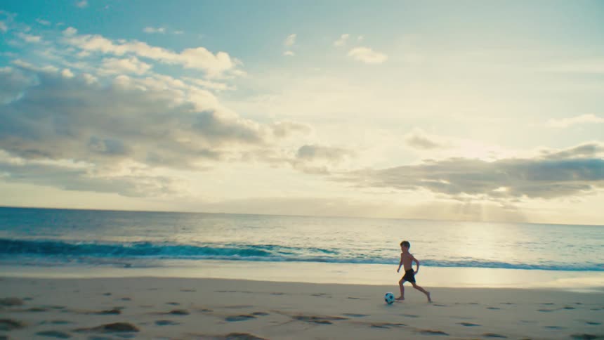Boy runs while kicking a soccer ball across the sand at the beach at sunset
