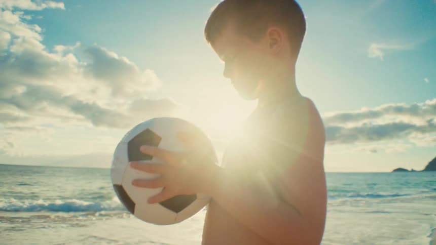 A young boy stands on a beach while holding a soccer ball