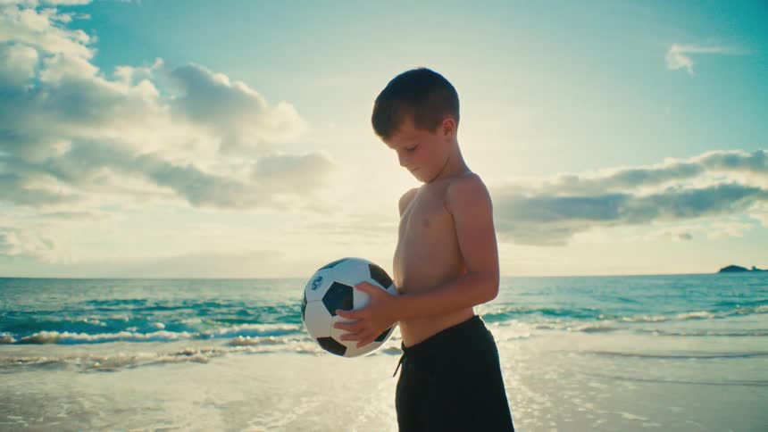 Boy standing on the beach holding a soccer ball as waves roll in to shore