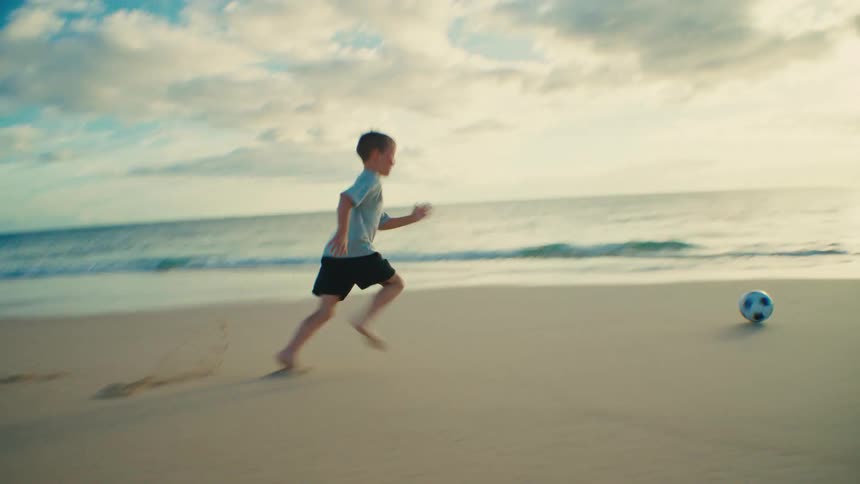 Boy running and kicking a soccer ball on the beach as waves roll in to shore