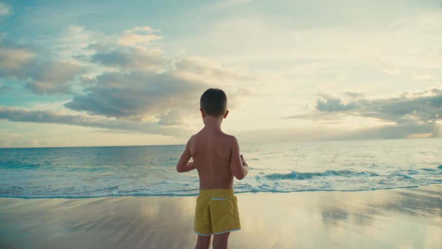 Boy without a shirt and wearing yellow shorts holding a ball while standing on the sand by the sea at the beach