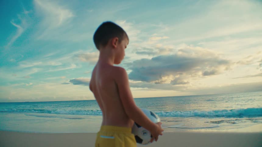 Close up of a boy holding a soccer ball while standing at the beach at sunset
