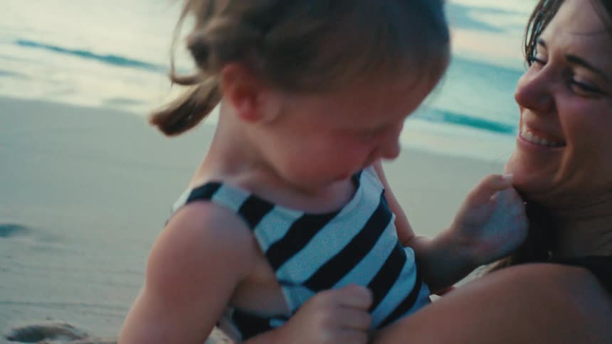 Mother kisses daughter on the cheek and both laughs while sitting on the beach at sunset
