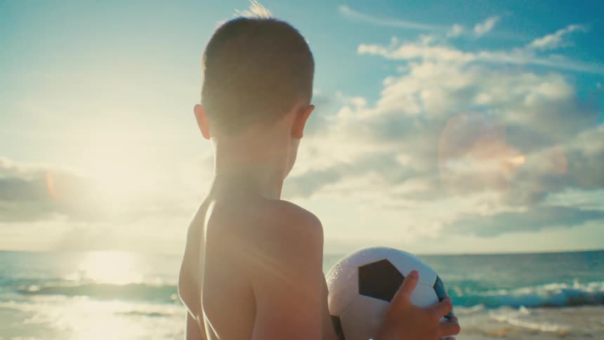 Close up of a boy holding a soccer ball while standing outdoors at the beach during daytime
