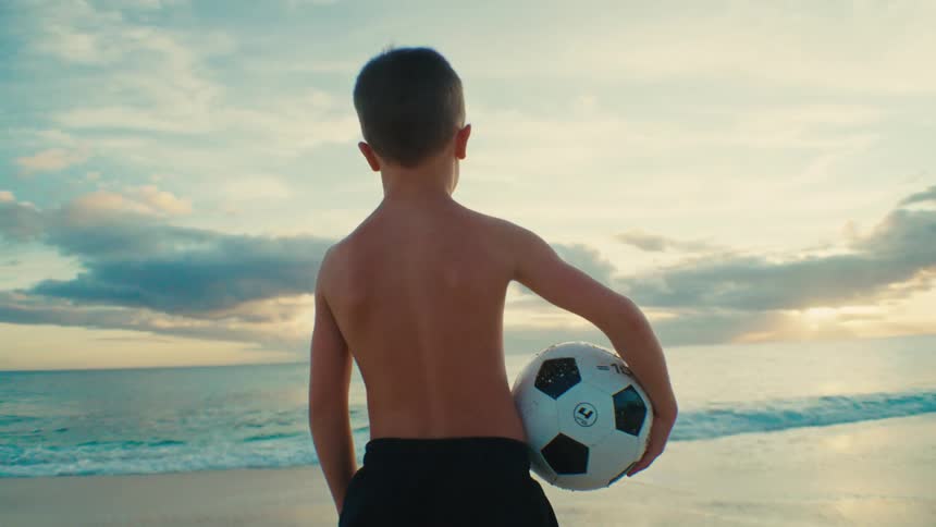 Boy standing on the beach holding a soccer ball as waves roll in to shore