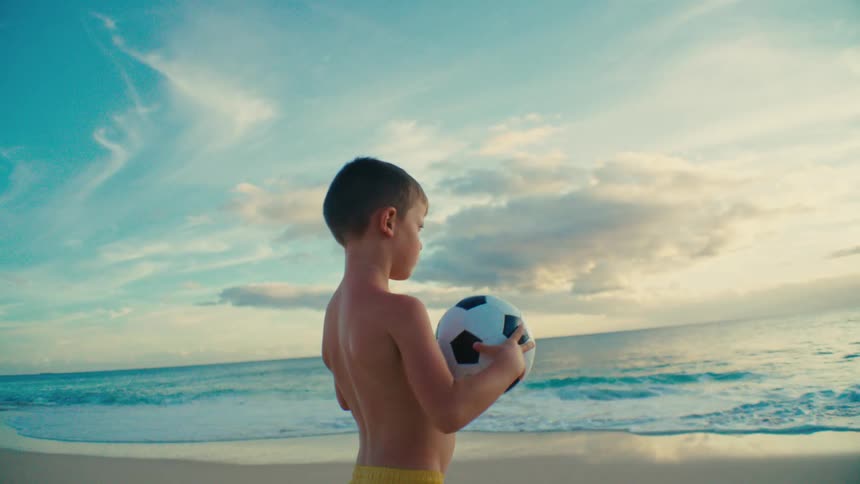 Little boy holding a soccer ball on the beach sand during the day
