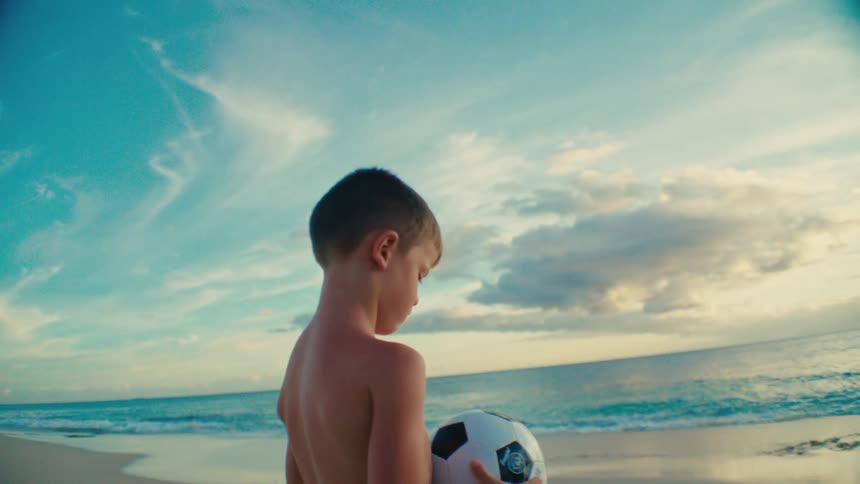A young boy with a soccer ball stands on the beach gazing at the ocean