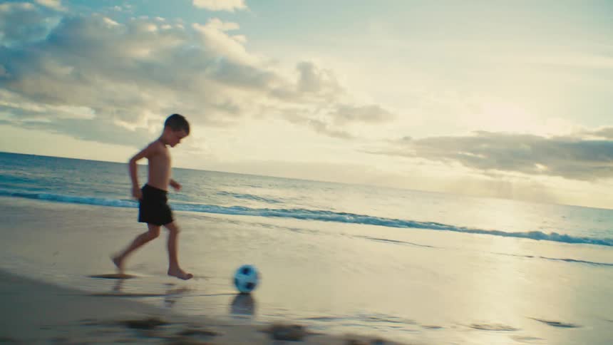 Young boy runs while kicking a soccer ball along the beach at sunset