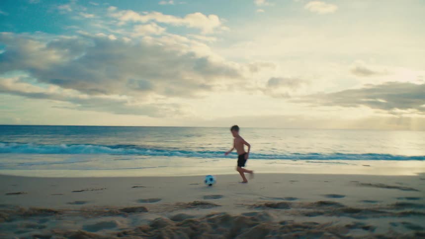 Boy kicks and runs after a soccer ball while playing at the beach at sunset
