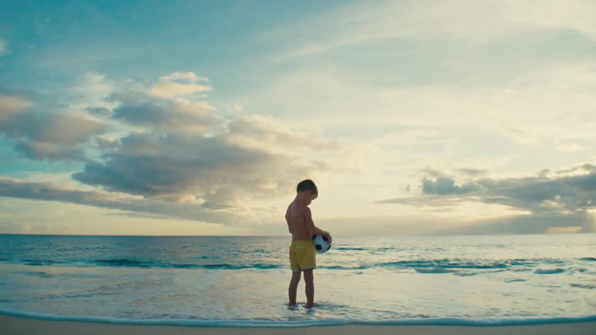 A young child holding a soccer ball stands on the beach as the waves wash over his feet at midday