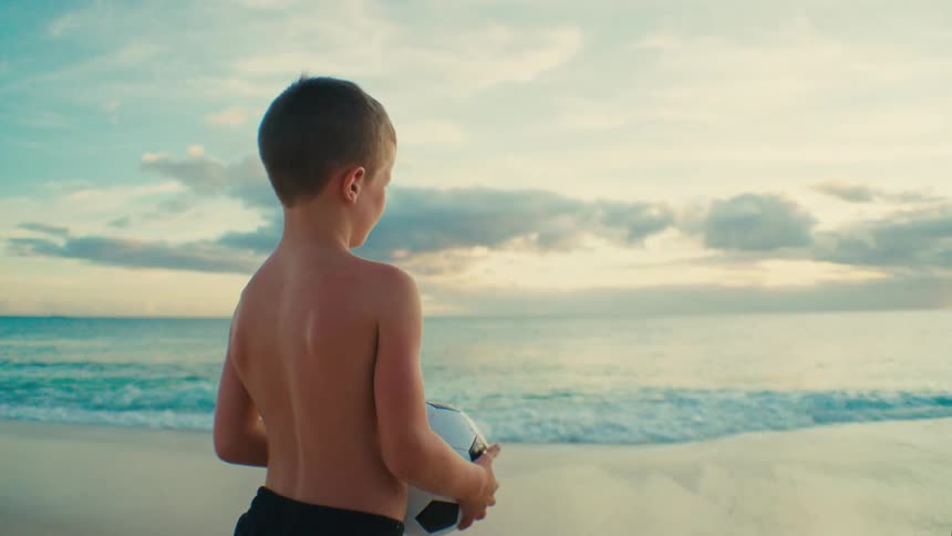 A child stands on a beach holding a soccer ball facing the ocean during sunset