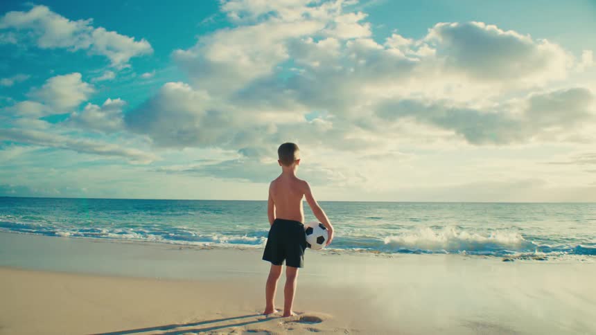 Boy standing on the beach holding a soccer ball watching waves roll in to shore