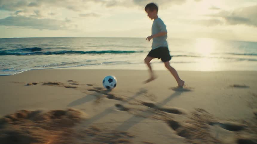 A boy kicks and chases after a soccer ball across the beach sand outdoors during daytime