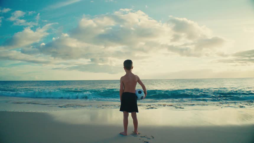 A child stands on a sandy beach holding a soccer ball facing the ocean under a sky filled with scattered clouds