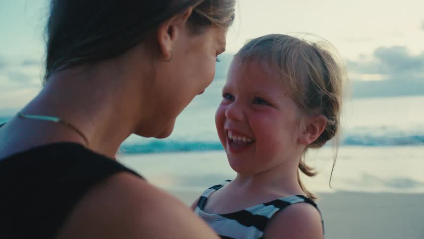 Mother lovingly embraces a smiling daughter on the beach at sunset