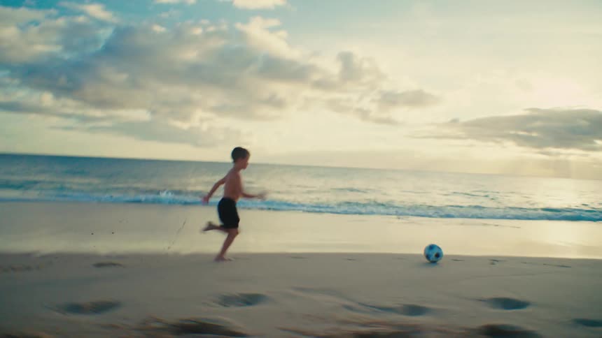 A boy kicks and chases after a soccer ball while playing at the beach at sunset