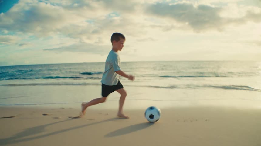 A young boy plays soccer alone on the beach at sunset