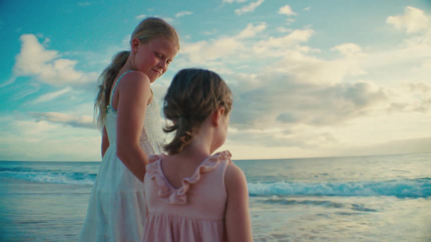 Two young girls stand smiling along a beach under a bright blue sky