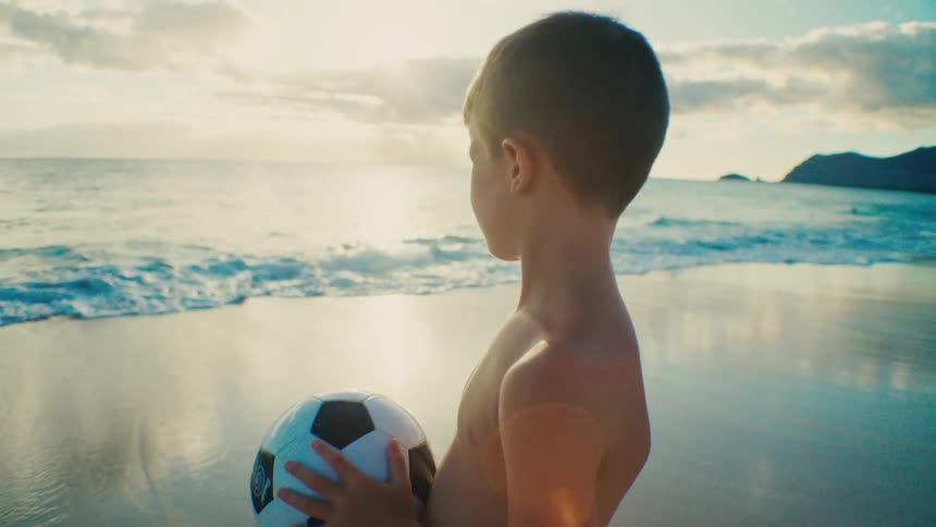 A young boy holds a soccer ball on a sandy beach near the ocean
