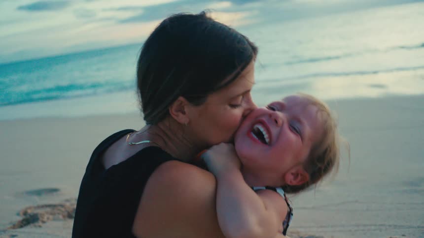 A woman affectionately hugs and kisses a laughing child on a beach at sunset