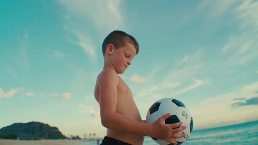 The child is holding a soccer ball on the beach