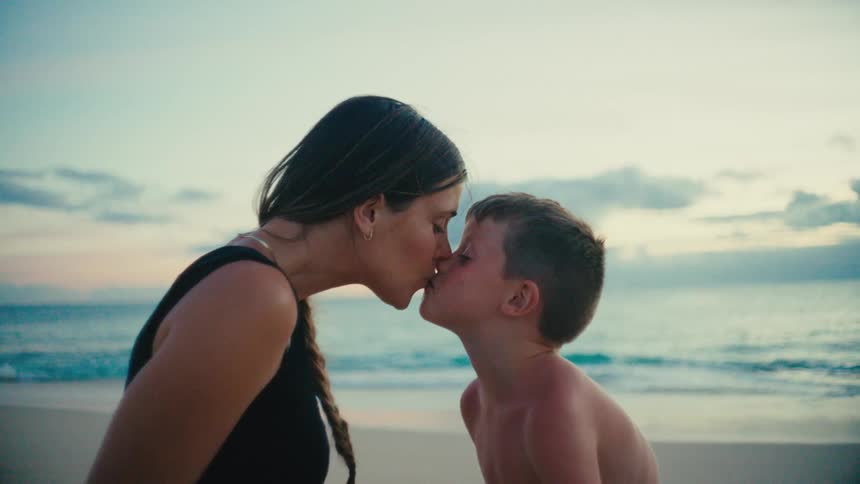 A mother kisses her child as they both sit on the beach sand at midday enjoying their time together