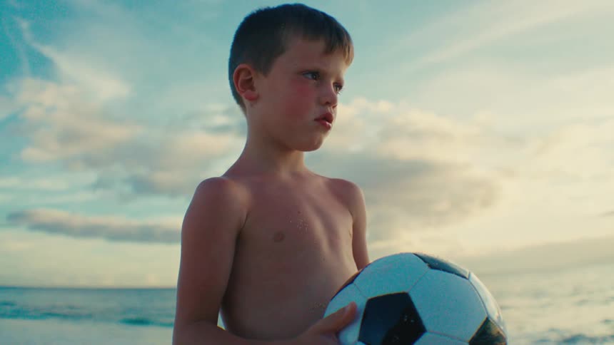 Young boy holding a soccer ball standing next to the ocean at dusk