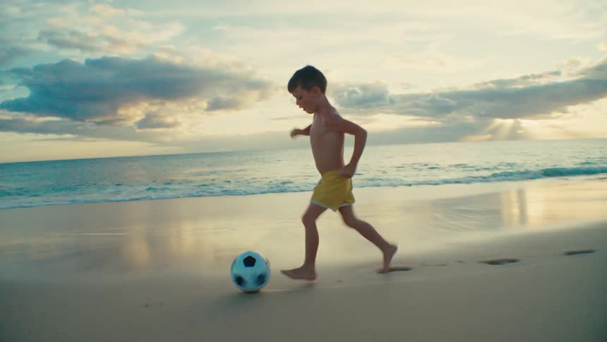 A young boy is playing soccer alone on a beach at sunset