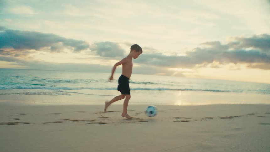 A boy kicks and runs after a soccer ball while playing at the beach during daytime