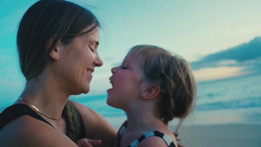 Mother smiles and kisses daughter while sitting outdoors at the beach during daytime