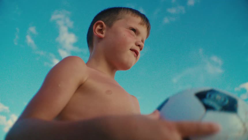 Sunlight shines upon a boy holding a soccer ball and turning it in his hands as he thinks