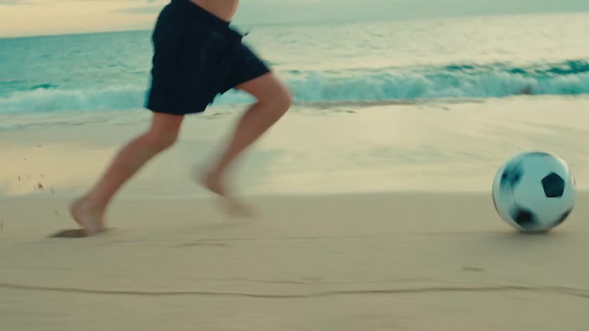 A boy is playing soccer barefoot on a sandy beach near the ocean